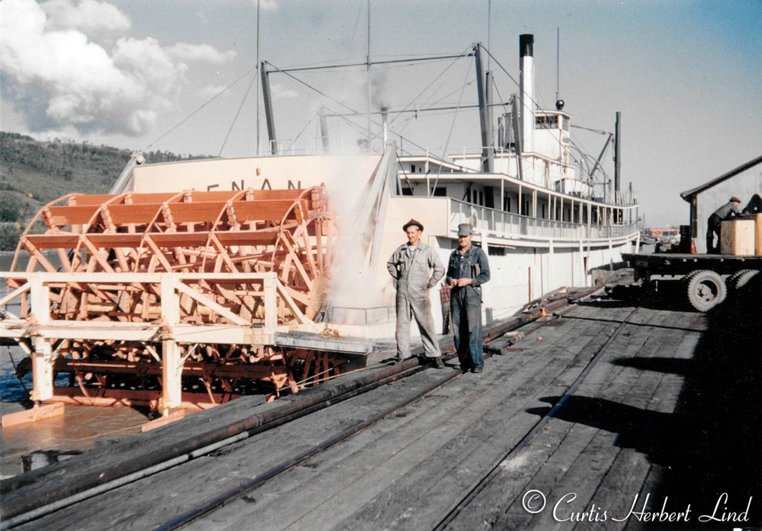 The Alaska Railroad riverboat NENANA at the town of Nenana facing up stream on the Tanana River. If you look close you can see the approach to the Mears Memorial bridge just off the bow of the boat. Looks like it is getting a new coat of red lead paint after about 50 years in service. Shadows are coming from due west indicating this is the height of summer and probably between 8 and 10 pm. The pipe down the edge of the dock was for delivering fuel oil to barges and boats. I believe the Nenana had been converted to oil by 1950. Curt Lind at left.